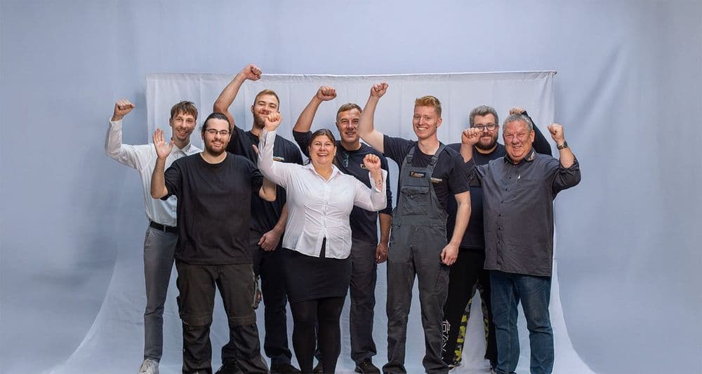 group picture 8 car dealership employees against a grey background raise their arms in the air in jubilation